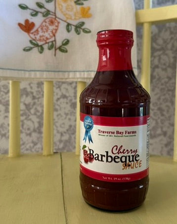 Bottle of Traverse Bay Farms Cherry Barbeque Sauce on a wooden surface with a patterned chair in the background.