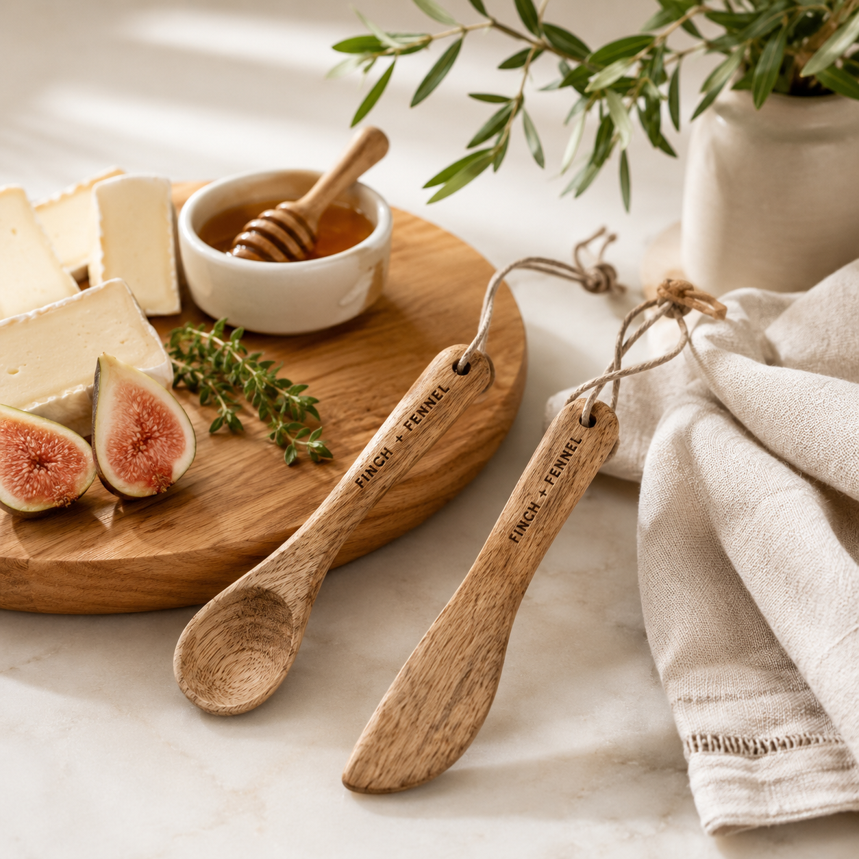 Wooden spoons with 'Fench & Fernel' branding on a table with cheese, figs, and honey.